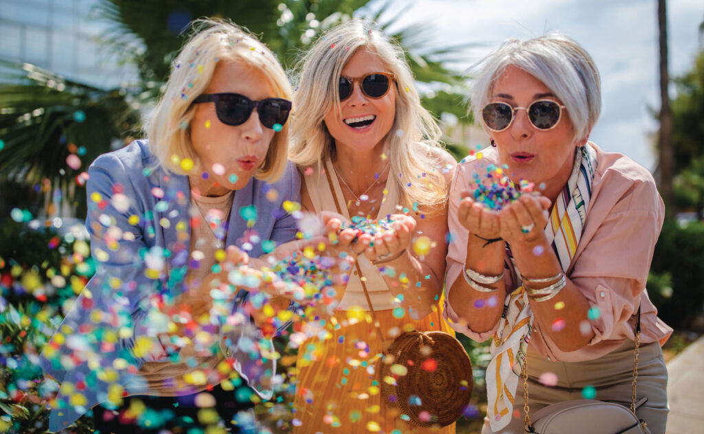 Three senior women wearing sunglasses cup their hands and blow colorful confetti at the camera with with smiles on their faces.